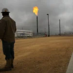 Person in hard hat observes a flare stack emitting fire at an industrial site under cloudy skies.