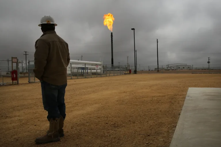 Person in hard hat observes a flare stack emitting fire at an industrial site under cloudy skies.