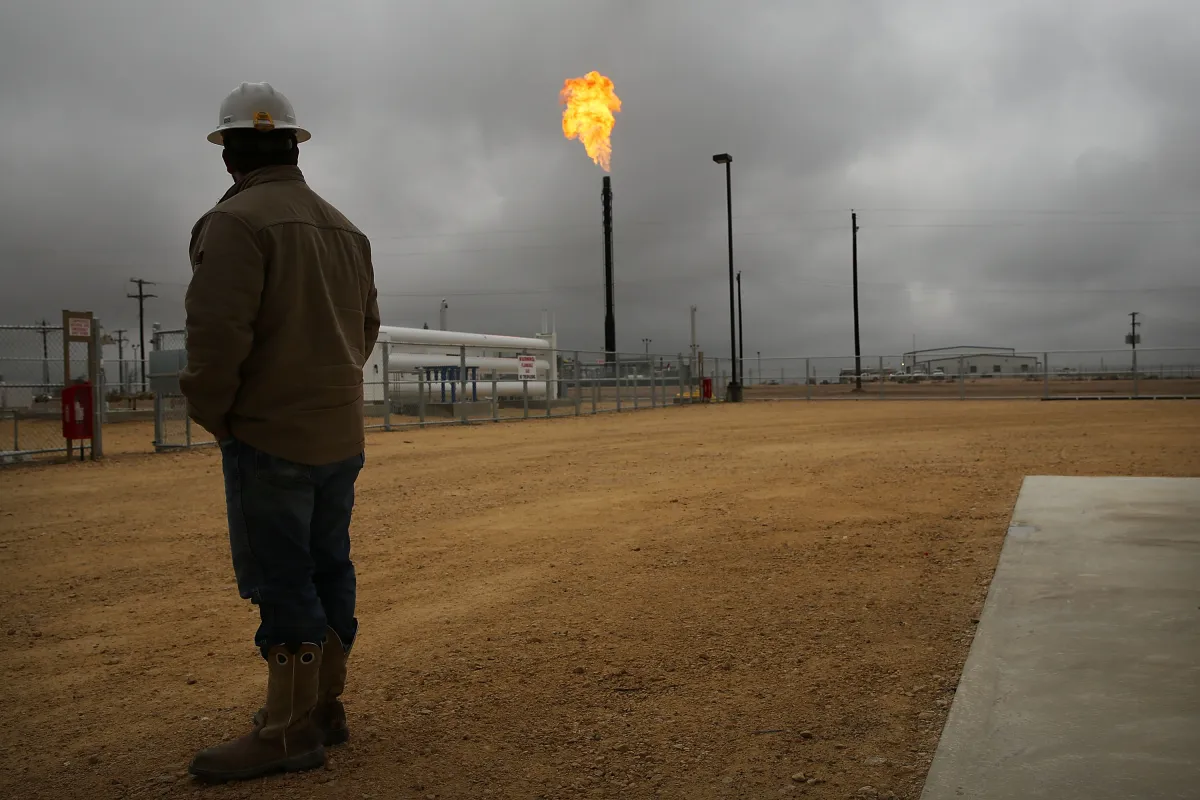 Person in hard hat observes a flare stack emitting fire at an industrial site under cloudy skies.