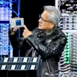 Person in a black leather jacket holds and points at a computer chip, with server racks in the background.