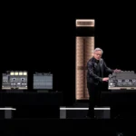 Man in a black jacket presenting computer hardware on stage with two large servers in the background.
