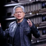 Person in a black leather jacket gestures while speaking, with server racks in the background.