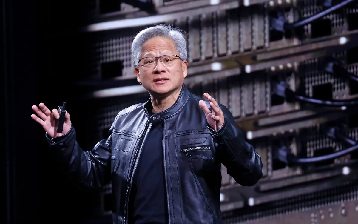Person in a black leather jacket gestures while speaking, with server racks in the background.