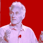 Man speaking with hands raised, wearing a light shirt and microphone, set against a red and blue background.
