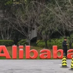 Large red Alibaba sign outdoors, person using phone nearby, white car parked, trees and bushes in the background.