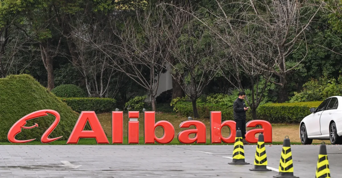 Large red Alibaba sign outdoors, person using phone nearby, white car parked, trees and bushes in the background.