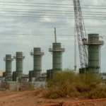 Five large industrial towers with a crane in a construction area, surrounded by fencing and vegetation under cloudy sky.