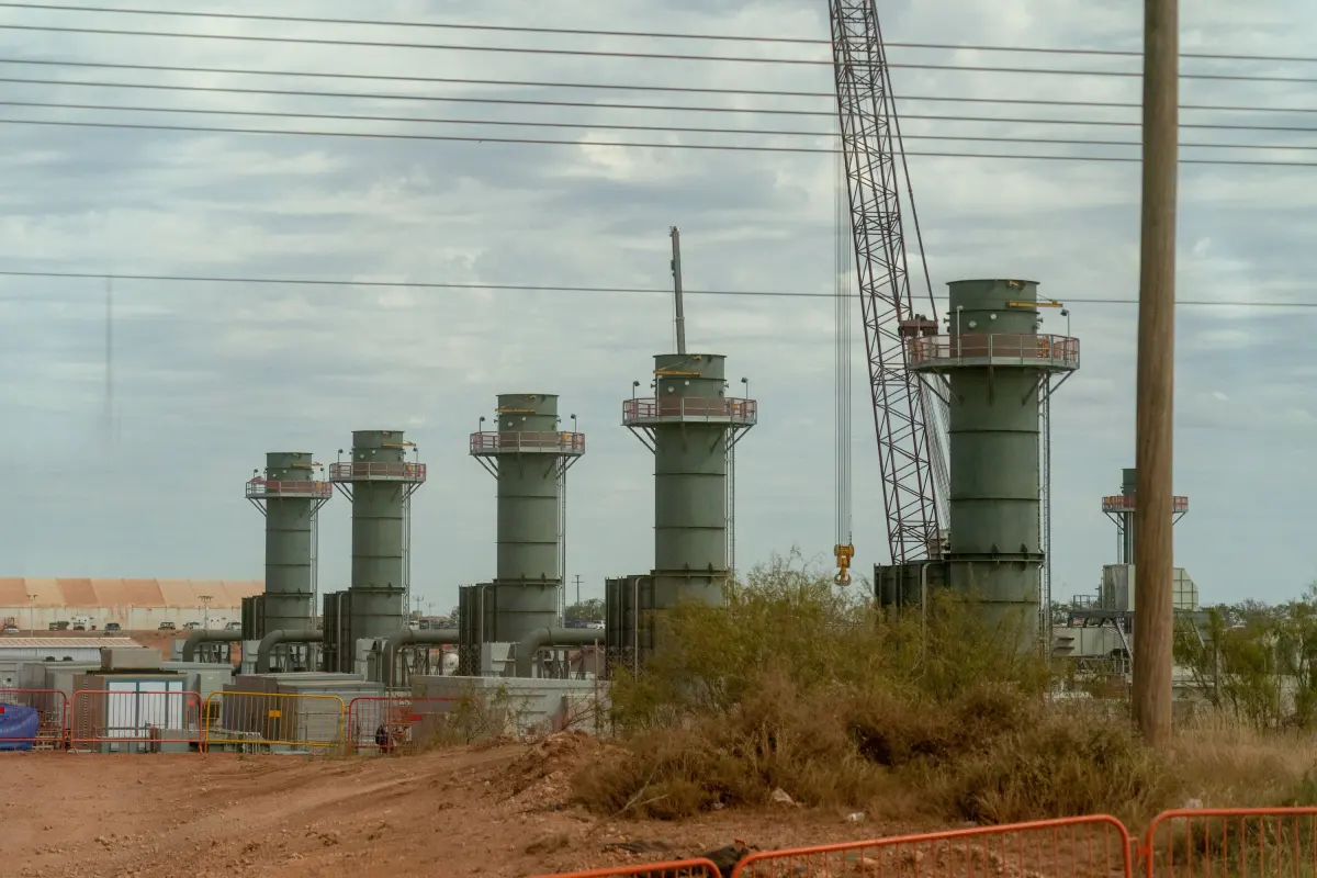 Five large industrial towers with a crane in a construction area, surrounded by fencing and vegetation under cloudy sky.