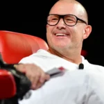 Person in white outfit smiling while sitting on a red chair, another person partially visible on the left.