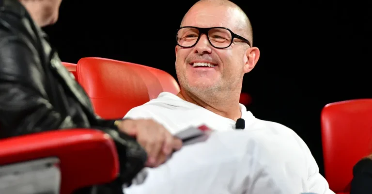 Person in white outfit smiling while sitting on a red chair, another person partially visible on the left.