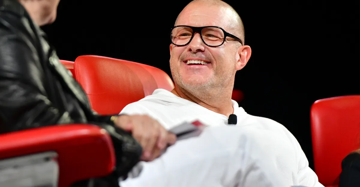 Person in white outfit smiling while sitting on a red chair, another person partially visible on the left.