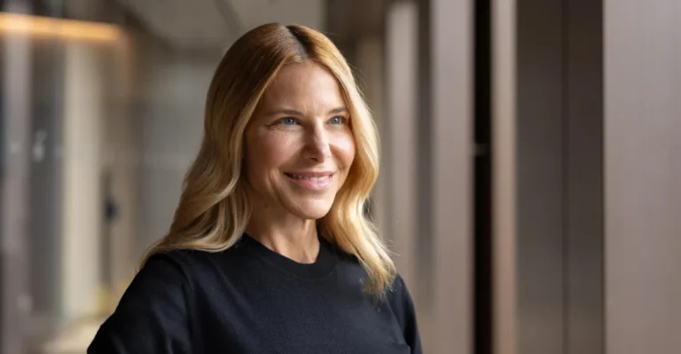 Smiling person with long blonde hair in a black shirt, standing indoors near a window with soft lighting.
