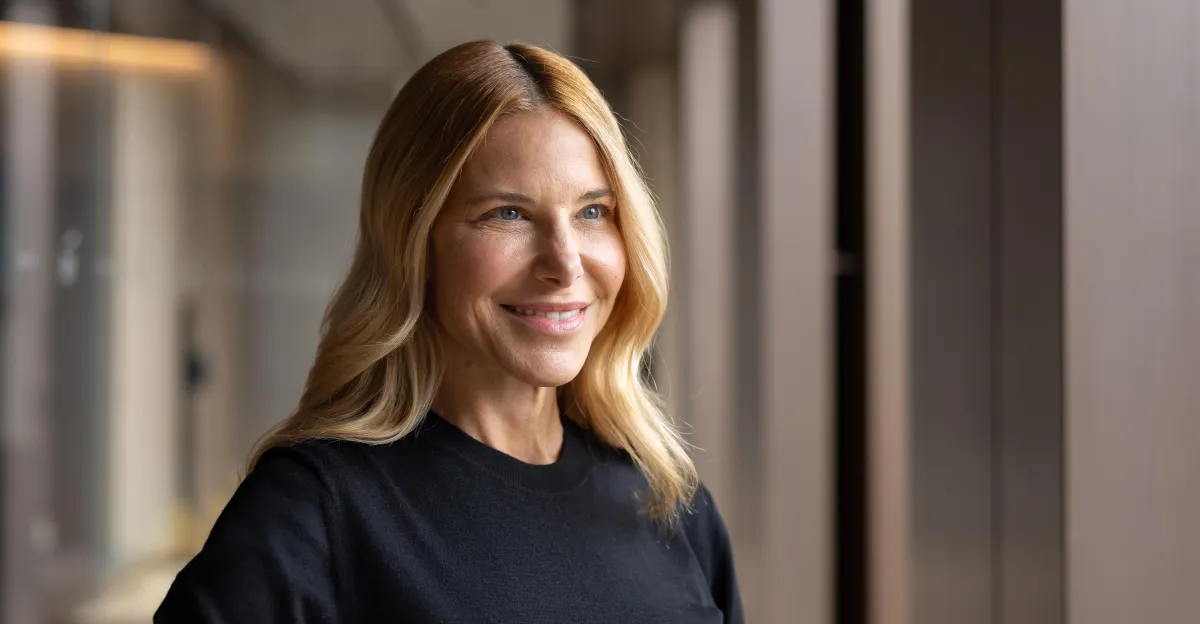 Smiling person with long blonde hair in a black shirt, standing indoors near a window with soft lighting.