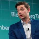 Person in blue suit speaking at Bloomberg Tech event, green backdrop with "Bloomberg Tech" text.