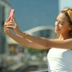 Woman taking a selfie with a smartphone outdoors, wearing a yellow headband and white top, cityscape in the background.