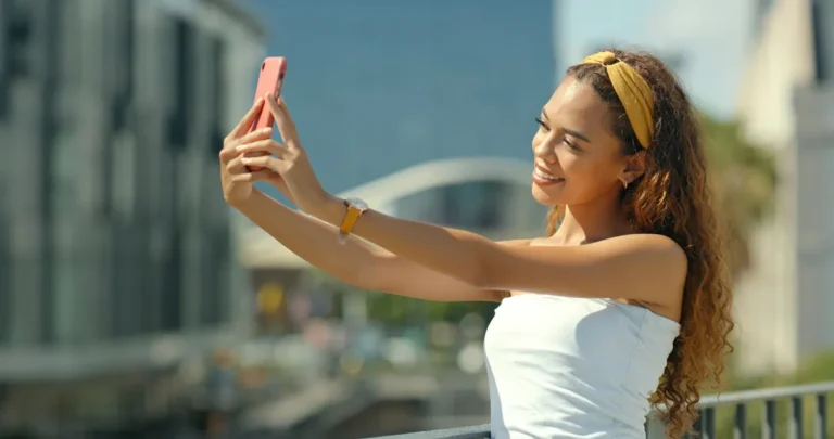 Woman taking a selfie with a smartphone outdoors, wearing a yellow headband and white top, cityscape in the background.