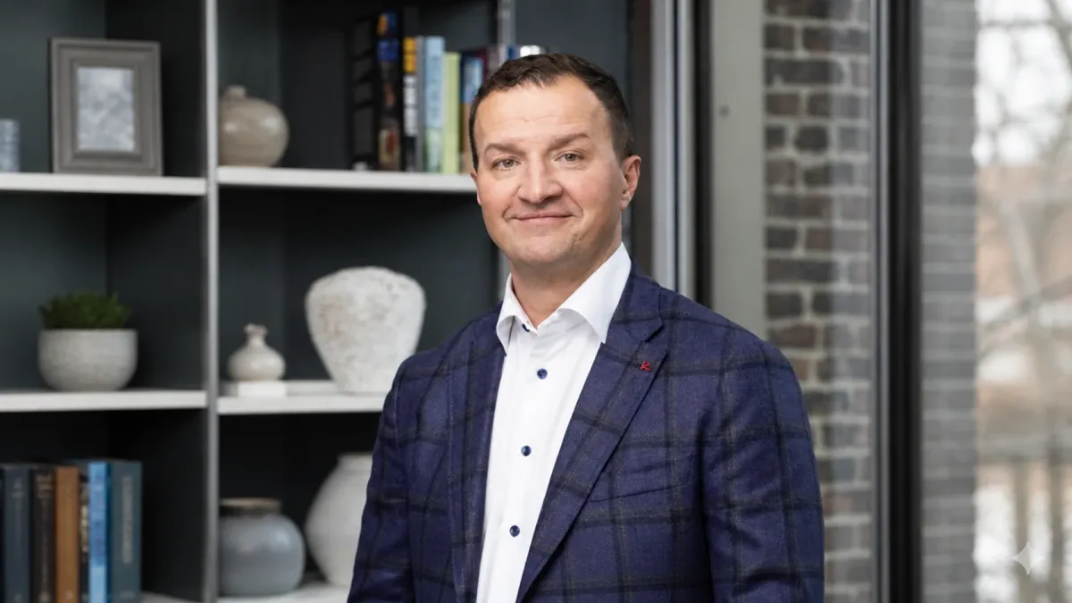 Man in a blue plaid suit stands in front of a bookshelf with decorative items and books, brick wall in the background.
