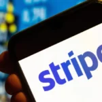 Smartphone displaying the Stripe logo, held up against a blurred background of blue and yellow numbers.