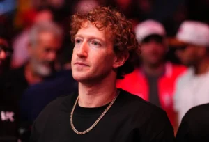 Man with curly hair and gold chain in a dark shirt, surrounded by a blurred crowd, looking intently forward.