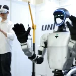 A humanoid robot with glowing faceplate raises its arms, while a person in VR gear operates controllers in a booth setting.
