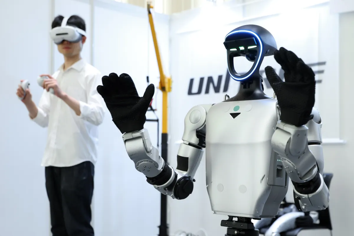 A humanoid robot with glowing faceplate raises its arms, while a person in VR gear operates controllers in a booth setting.