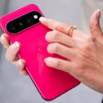 Person holding a bright pink smartphone, focusing on the camera module with a finger, wearing gold rings.