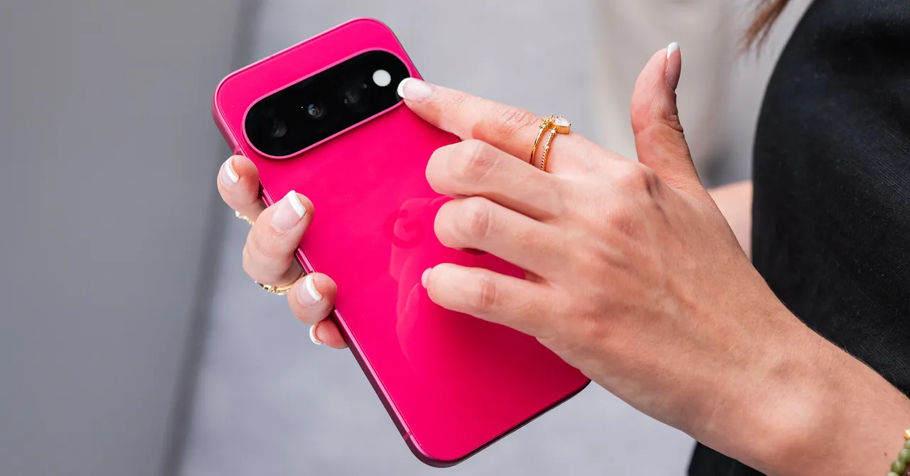 Person holding a bright pink smartphone, focusing on the camera module with a finger, wearing gold rings.