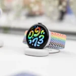 Smartwatch with colorful woven band on a charging stand, displaying vibrant numbers; earbuds case in the background.