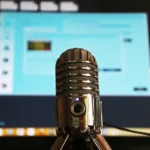A silver microphone in focus with a blurred computer screen displaying a blue interface in the background.
