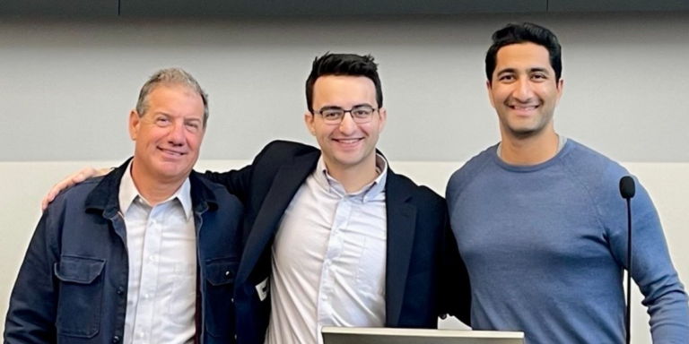 Three men standing together and smiling, one wearing a suit and glasses, the others in casual clothing, with a microphone...