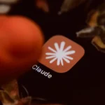 Finger touching a smartphone screen displaying an app icon labeled "Claude" with a starburst design.