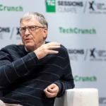 Person speaking on stage at TechCrunch event, wearing glasses and a striped sweater, with a microphone on their collar.