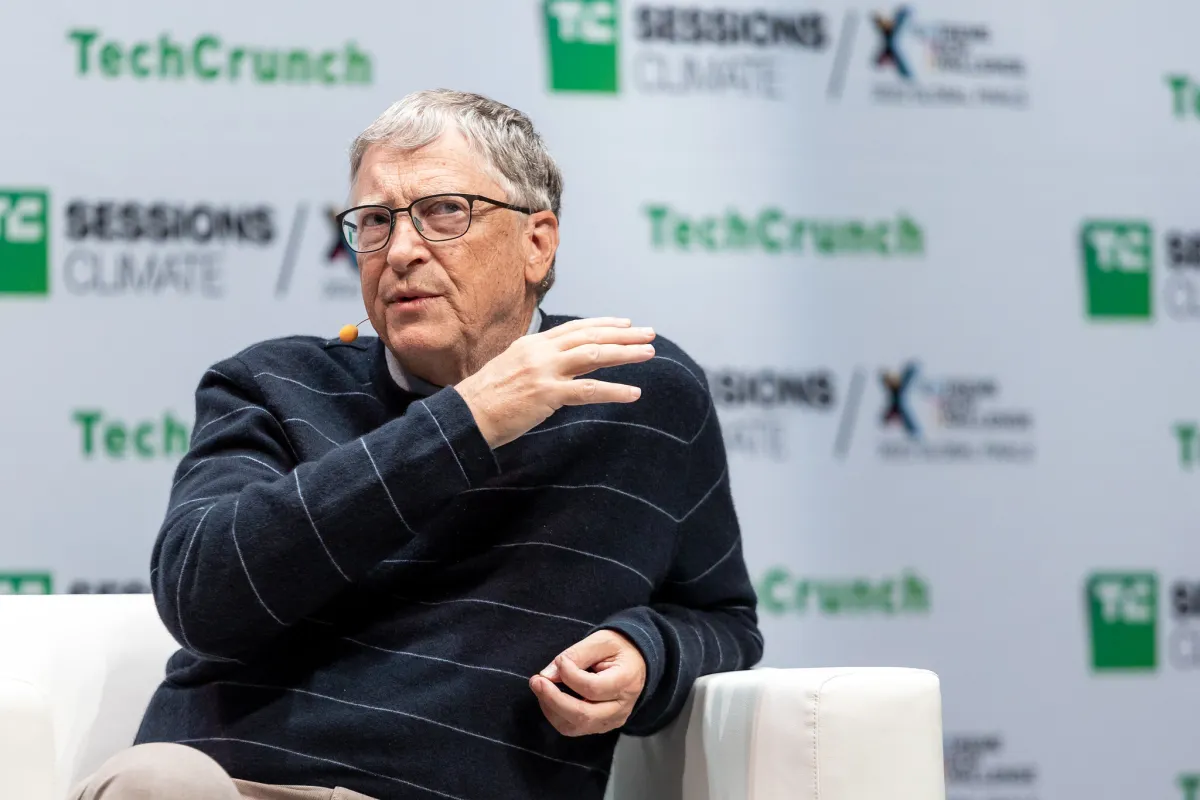 Person speaking on stage at TechCrunch event, wearing glasses and a striped sweater, with a microphone on their collar.