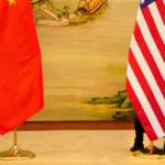 Chinese and American flags on stands, person peeking from behind the American flag, mural of traditional scene in background.