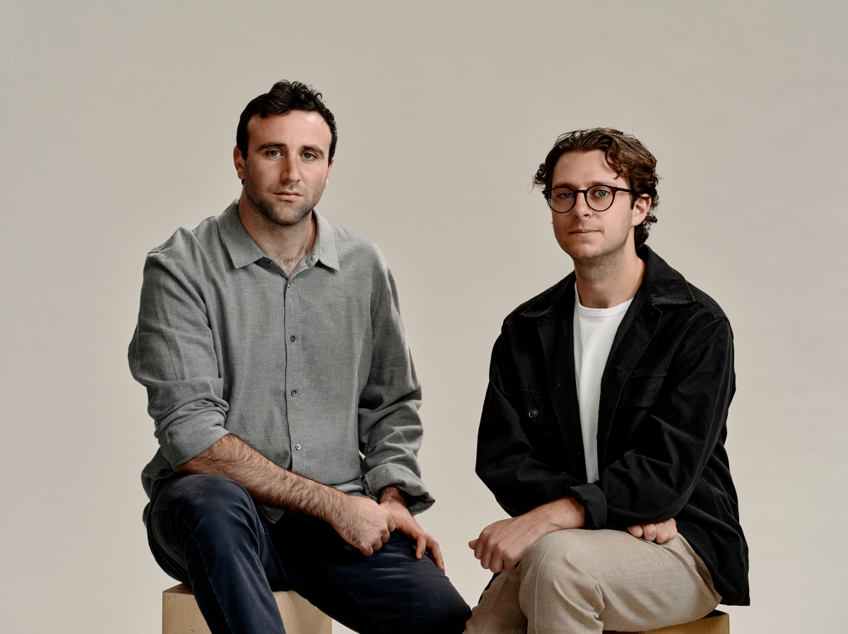 Two men seated, one wearing a gray shirt and the other a black jacket, against a neutral background.