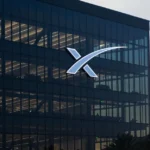 SpaceX logo on a glass building facade, with an American flag in the foreground and a cloudy sky in the background.
