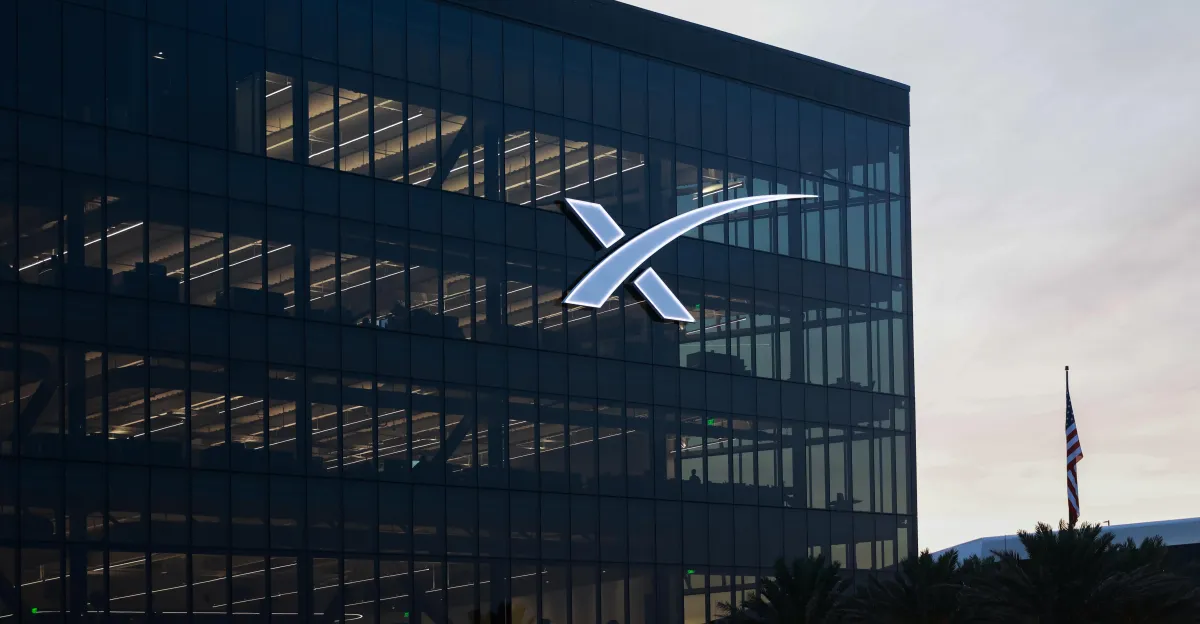 SpaceX logo on a glass building facade, with an American flag in the foreground and a cloudy sky in the background.