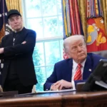 Two men in the Oval Office, one sitting at a desk, the other standing with arms crossed, flags and military emblems in bac...