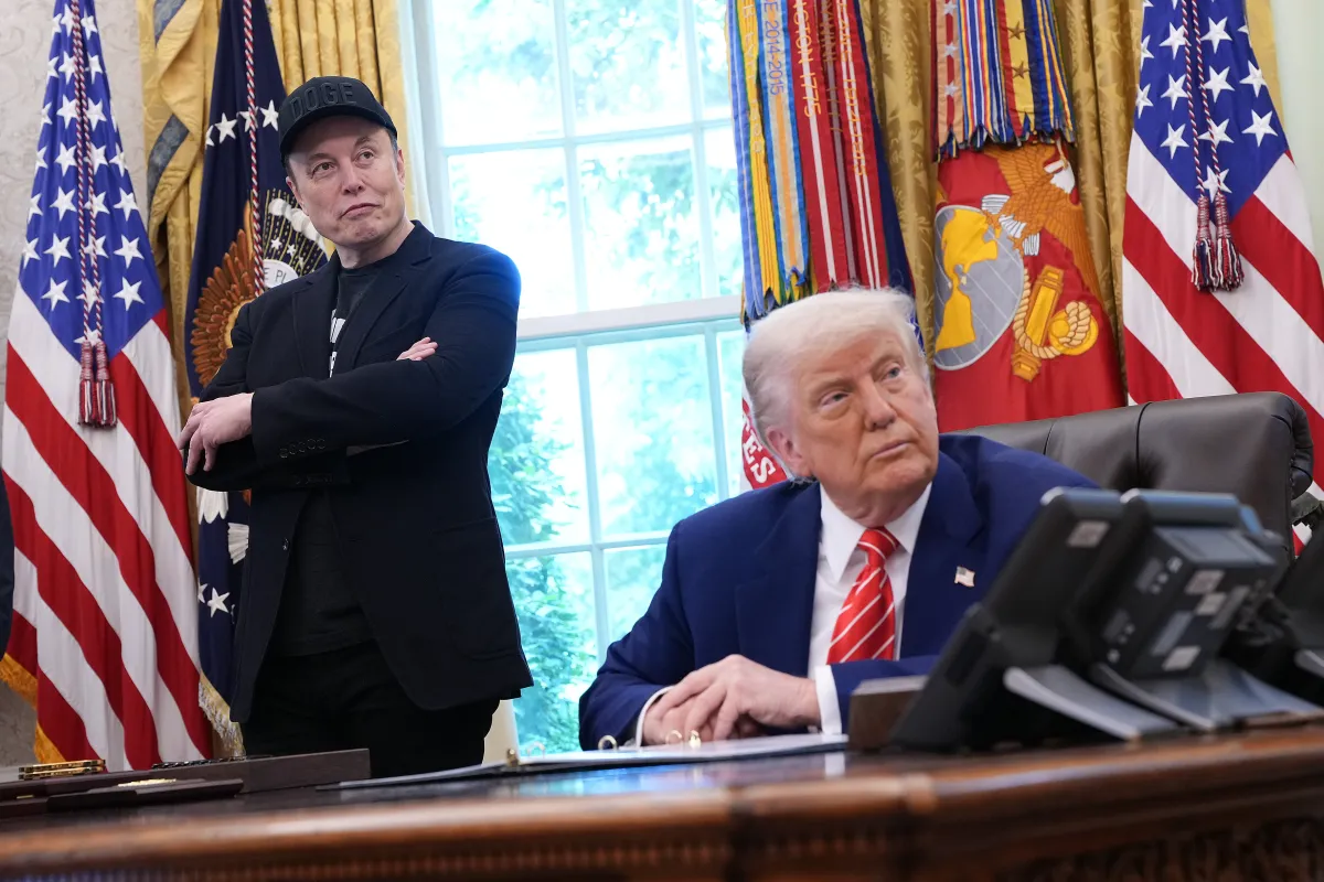 Two men in the Oval Office, one sitting at a desk, the other standing with arms crossed, flags and military emblems in bac...