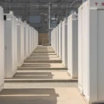 Rows of white Tesla battery units in an outdoor installation, with the Tesla logo visible on the rightmost unit.