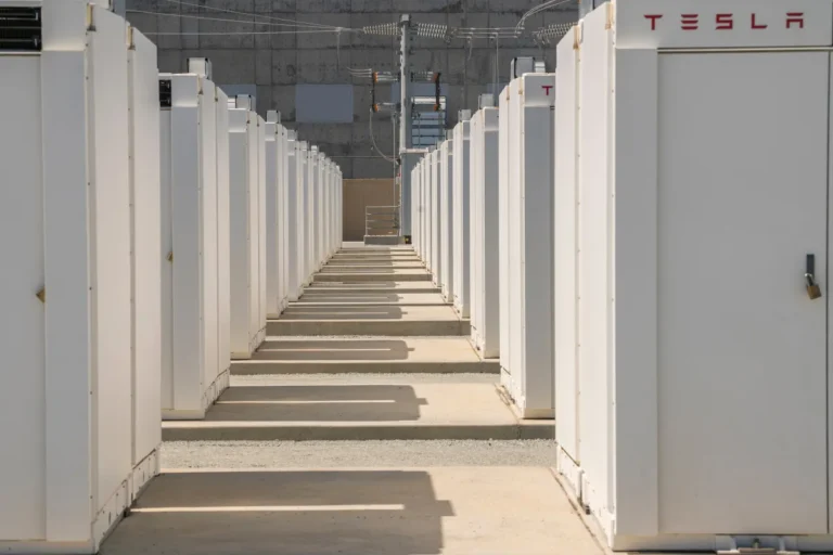 Rows of white Tesla battery units in an outdoor installation, with the Tesla logo visible on the rightmost unit.