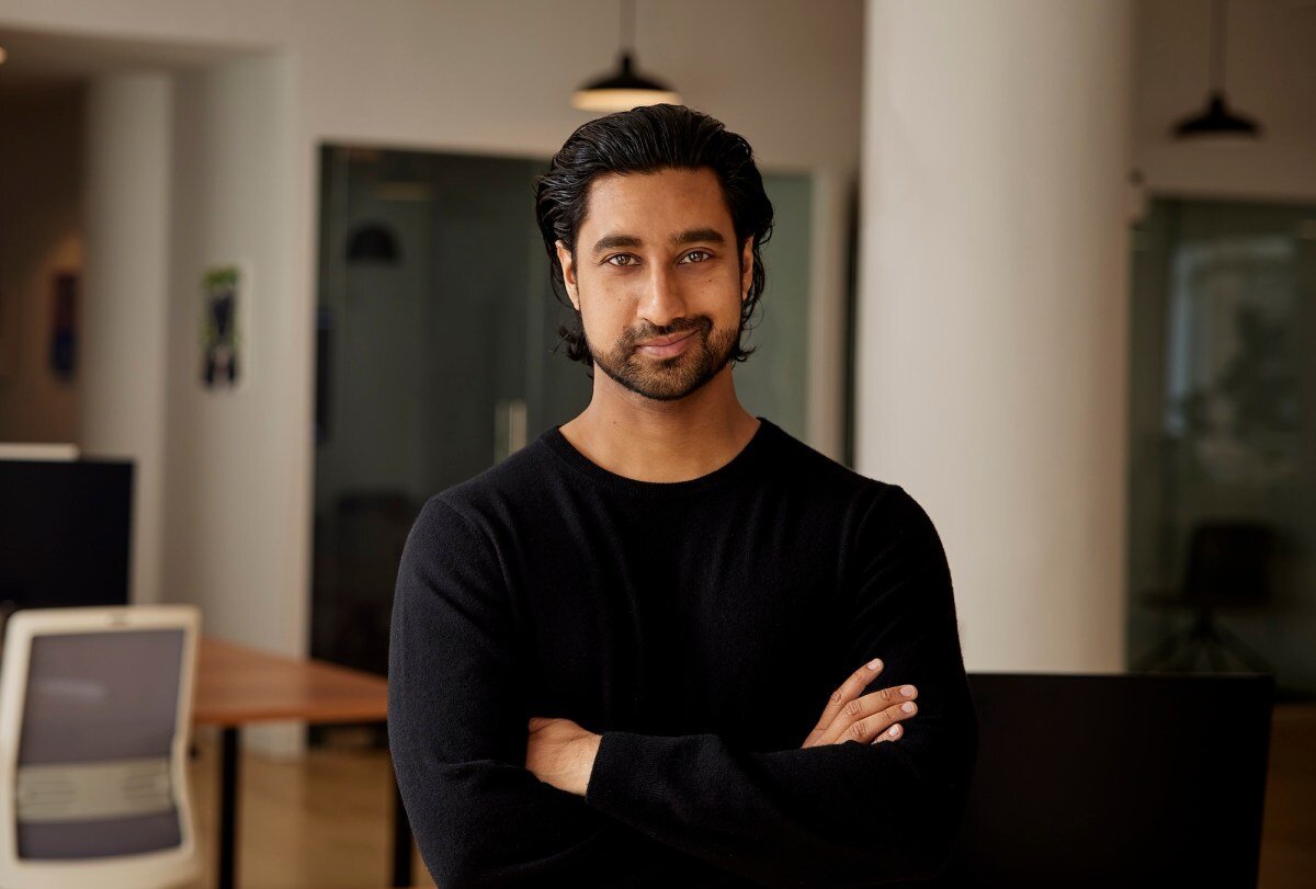 Person with dark hair and beard in a black sweater, standing with arms crossed in a modern office setting.