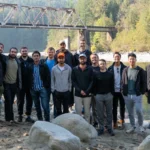 Group of 14 people standing on rocky riverbank with a rusty truss bridge and forest in the background.