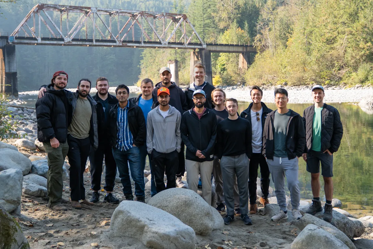 Group of 14 people standing on rocky riverbank with a rusty truss bridge and forest in the background.