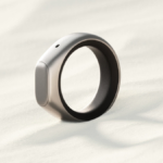 A sleek, modern ring with a metallic finish on a textured, light-colored surface, casting a soft shadow.