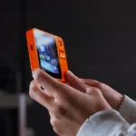 Hands holding an orange device with a screen displaying an interface, dark blurred background