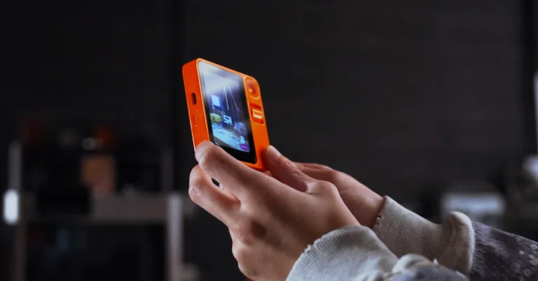 Hands holding an orange device with a screen displaying an interface, dark blurred background