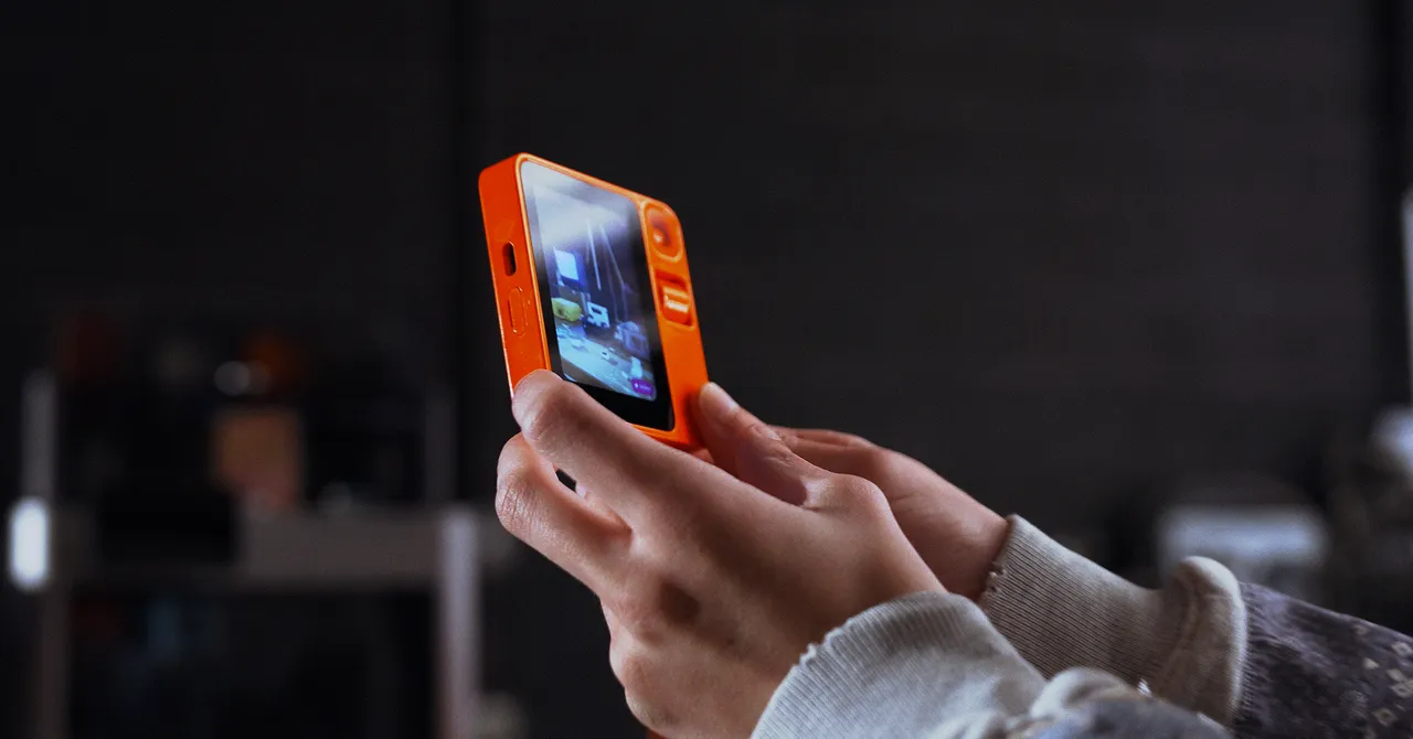 Hands holding an orange device with a screen displaying an interface, dark blurred background