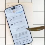Smartphone displaying shopping list app on a grid-patterned surface with a to-do list and plant leaves nearby.