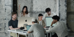 Six people collaborating at a table with laptops and computers in a modern office setting.